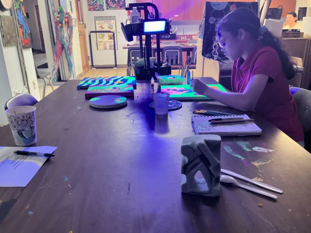 A colorful workspace featuring a wooden table cluttered with art supplies, glowing neon artwork, and a girl engaged in creative tasks.