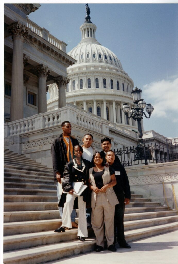 An adult teacher guides three young children, who have dreadlocks, as they write names and corresponding colors on a small whiteboard during a classroom activity